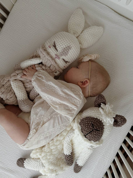  baby laying on a bed next to a stuffed bunny and alpaca  toys in neutral colours.