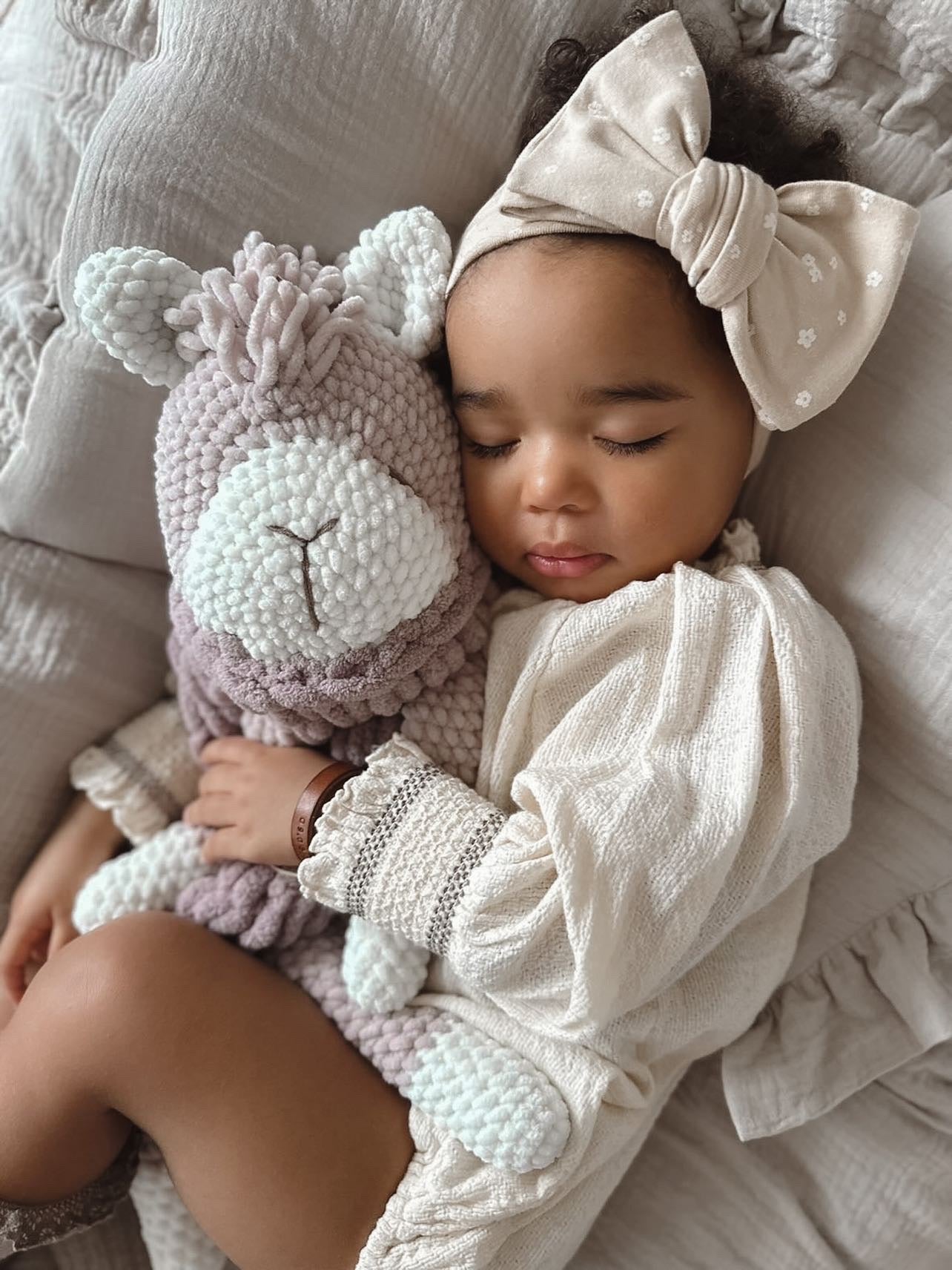  baby laying on a bed next to a stuffed alpaca toy in neutral colours.