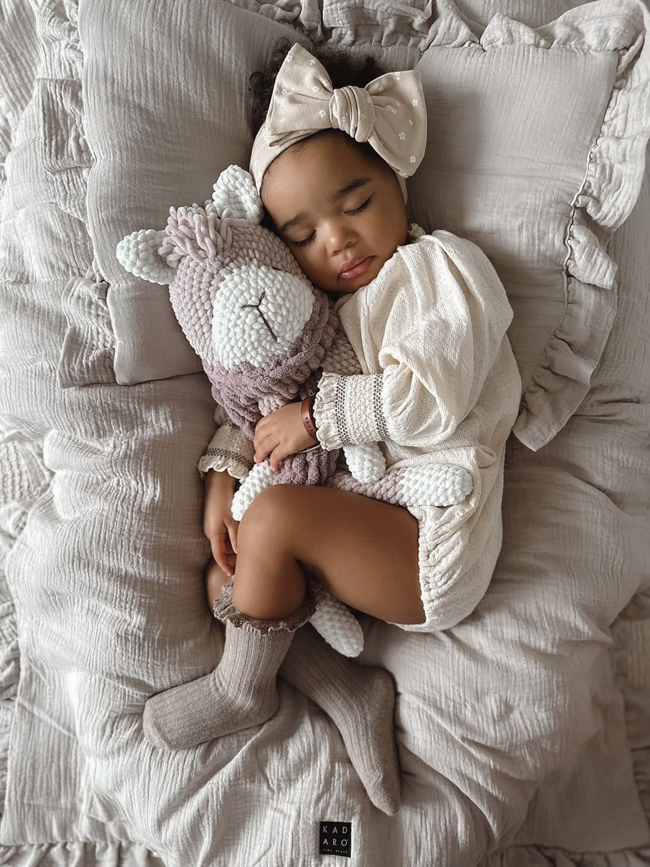 a baby laying on a bed next to a stuffed alpaca toy in neutral colours.