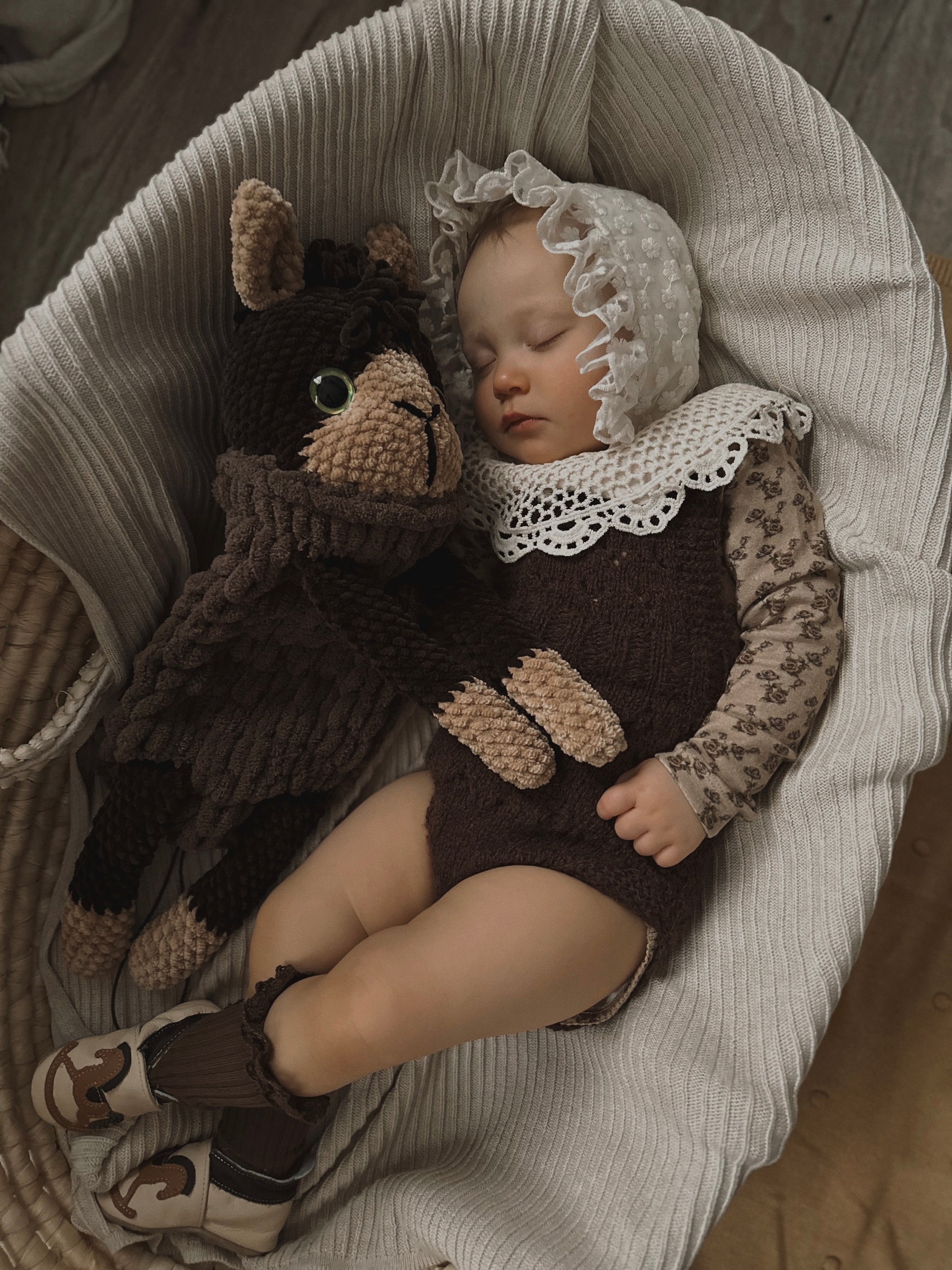  baby laying on a bed next to a stuffed alpaca toy in brown colours.